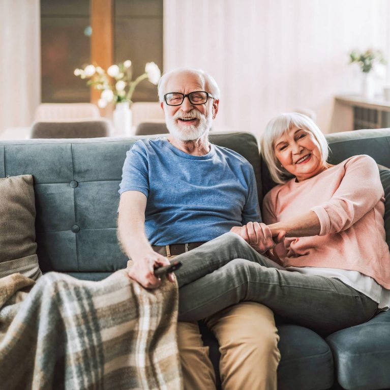 Ein älterer Mann und eine ältere Frau sitzen auf dem Sofa und lächeln. Beide haben weiße Haare und halten Händchen. Der Mann hält eine TV-Fernbedienung in der Hand.