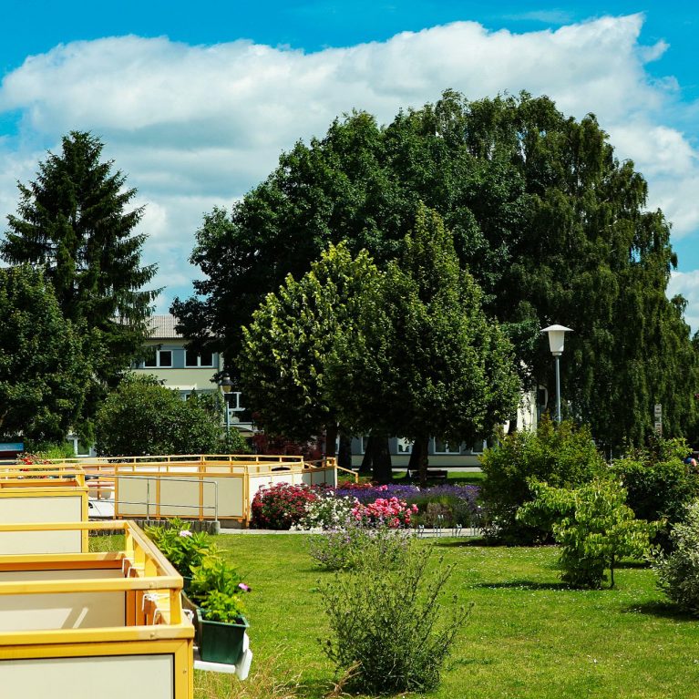Eine parkähnliche Anlage mit Balkonen von Häusern und eine Terrasse. Mehrere grüne Bäume und Büsche stehen in der Grünanlage.